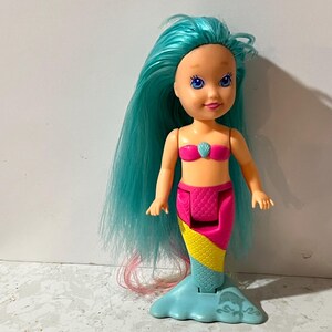 May include: A plastic mermaid doll with long, teal hair and a pink seashell top. The doll has a pink and yellow tail with a blue fin base. The doll's face has blue eyes and a smiling expression.