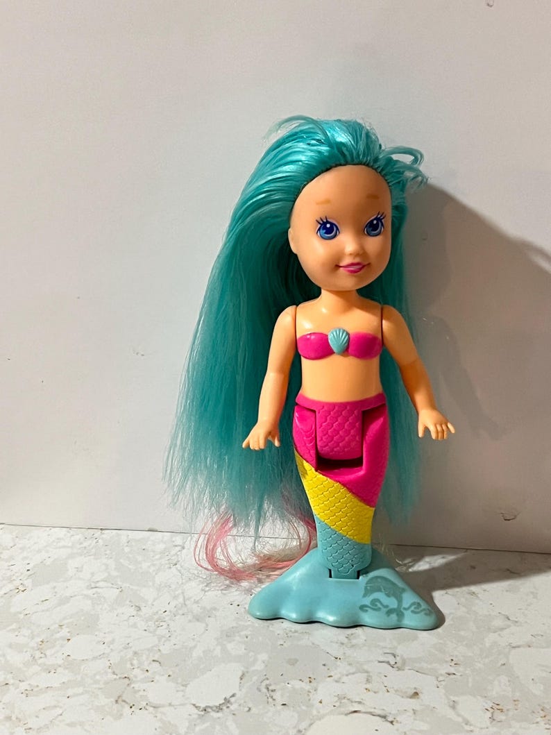 May include: A plastic mermaid doll with long, teal hair and a pink seashell top. The doll has a pink and yellow tail with a blue fin base. The doll's face has blue eyes and a smiling expression.