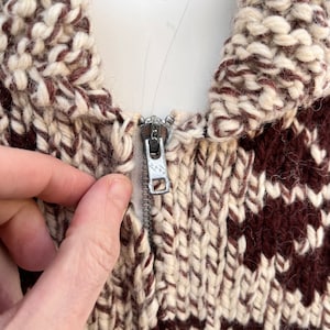 May include: Close-up of a brown and white knitted sweater with a zipper closure. The zipper is silver and has a small metal pull tab.