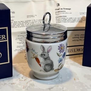 May include: A white ceramic egg coddler with a silver lid and a handle. The coddler features a whimsical illustration of a rabbit holding a carrot in a garden setting with flowers. The coddler is sitting on a countertop.