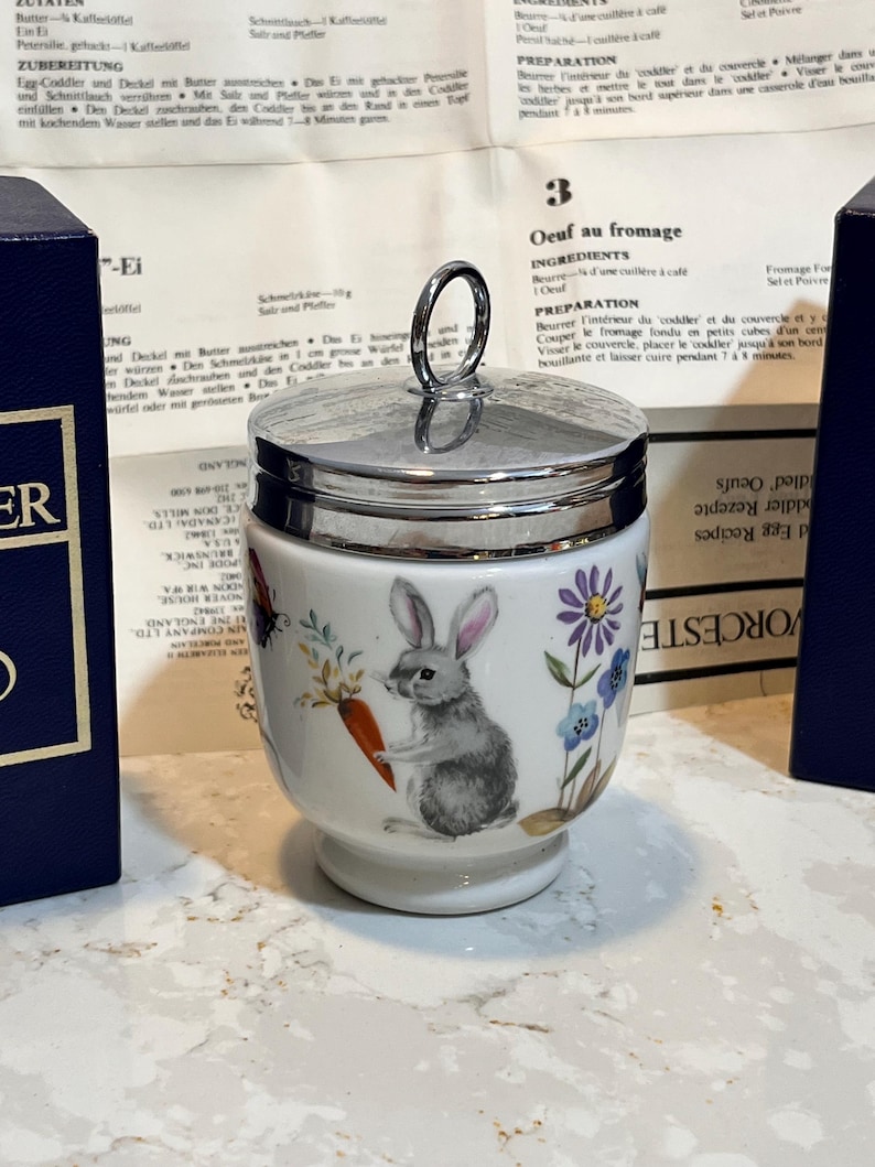 May include: A white ceramic egg coddler with a silver lid and a handle. The coddler features a whimsical illustration of a rabbit holding a carrot in a garden setting with flowers. The coddler is sitting on a countertop.
