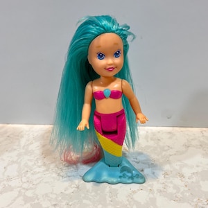 May include: A mermaid doll with turquoise hair and blue eyes. The doll wears a pink seashell top and a pink, yellow, and blue tail. The doll's tail is supported by a blue base with wave details.