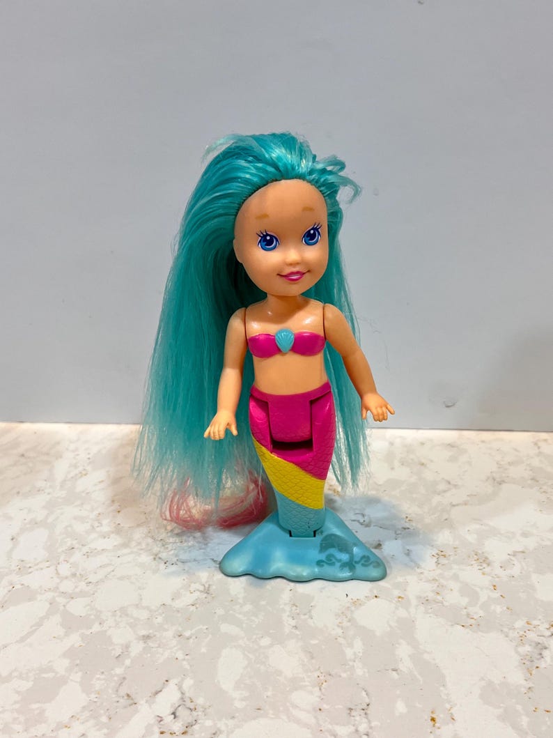 May include: A mermaid doll with turquoise hair and blue eyes. The doll wears a pink seashell top and a pink, yellow, and blue tail. The doll's tail is supported by a blue base with wave details.