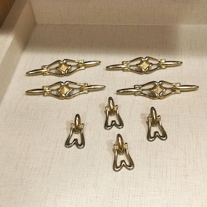 Op de afbeelding: Set van acht gouden kastbeslagknoppen. Vier zijn lange, rechthoekige knoppen met een decoratief ontwerp. Vier zijn kleinere, ronde knoppen met een decoratief ontwerp.