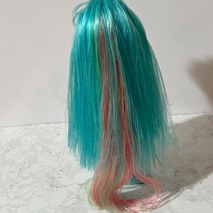 May include: A doll wig with vibrant turquoise hair, accented with streaks of pink, yellow, and blonde. The wig is styled with long, flowing strands, creating a colorful and playful appearance. The wig is likely for a doll or cosplay.
