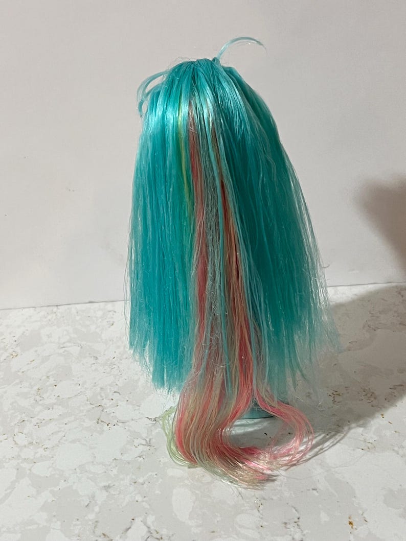 May include: A doll wig with vibrant turquoise hair, accented with streaks of pink, yellow, and blonde. The wig is styled with long, flowing strands, creating a colorful and playful appearance. The wig is likely for a doll or cosplay.