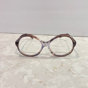 May include: Oval-shaped eyeglasses with a marbled brown and blue pattern. The frames have a unique, angular design at the temples and bridge. The lenses are clear, and the glasses are set against a neutral background.