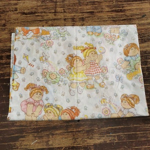 Pode incluir: Tecido branco com um padrão repetido de Cabbage Patch Kids brincando. As bonecas usam roupas coloridas e estão cercadas por flores, borboletas e arco-íris.