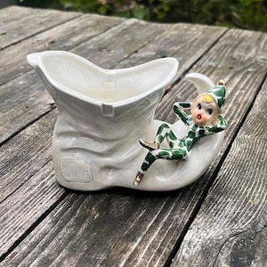 Könnte beinhalten: Ein weißer Keramik-Stiefel-Pflanztopf mit einer Elfenfigur. Der Elf in Grün und Weiß liegt auf einem Halbmond. Der Pflanztopf hat eine strukturierte Oberfläche und eine kleine Öffnung für Pflanzen oder Blumen.