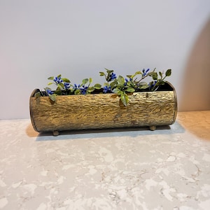 Peut inclure: Un pot de fleurs cylindrique doré avec une surface texturée, ressemblant à une bûche. Le pot est rempli de petites feuilles vertes et de fleurs bleues en forme de baies. Le pot repose sur trois petits pieds ronds.