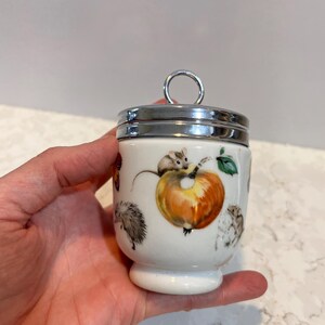 May include: White ceramic egg cup with a silver lid and a handle. The cup is decorated with a colorful illustration of a squirrel, a hedgehog, and an apple.
