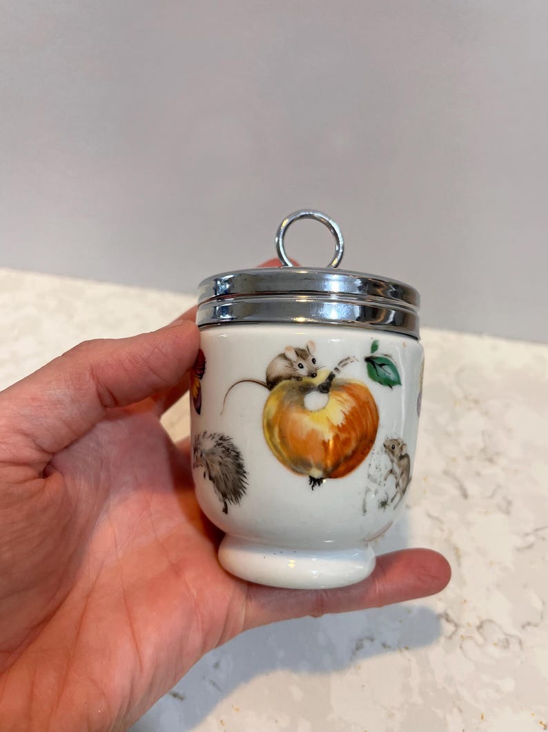 May include: White ceramic egg cup with a silver lid and a handle. The cup is decorated with a colorful illustration of a squirrel, a hedgehog, and an apple.