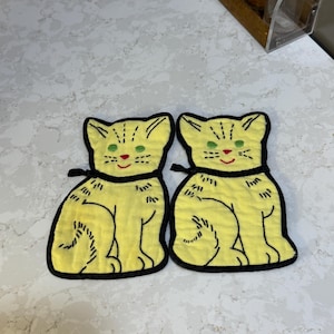 Peut inclure: Deux dessous de plat jaunes en forme de chat avec des contours et des détails noirs. Chaque chat a des yeux verts, un nez rouge et un collier noir. Les dessous de plat sont en tissu texturé.