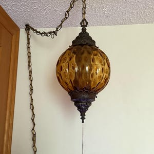 Peut inclure: Une lampe suspendue dotée d'un abat-jour en verre ambré de forme ronde avec une surface texturée et alvéolée. La lampe est suspendue par une chaîne en métal et comprend des garnitures en métal foncé ornementées en haut et en bas, avec une tirette.