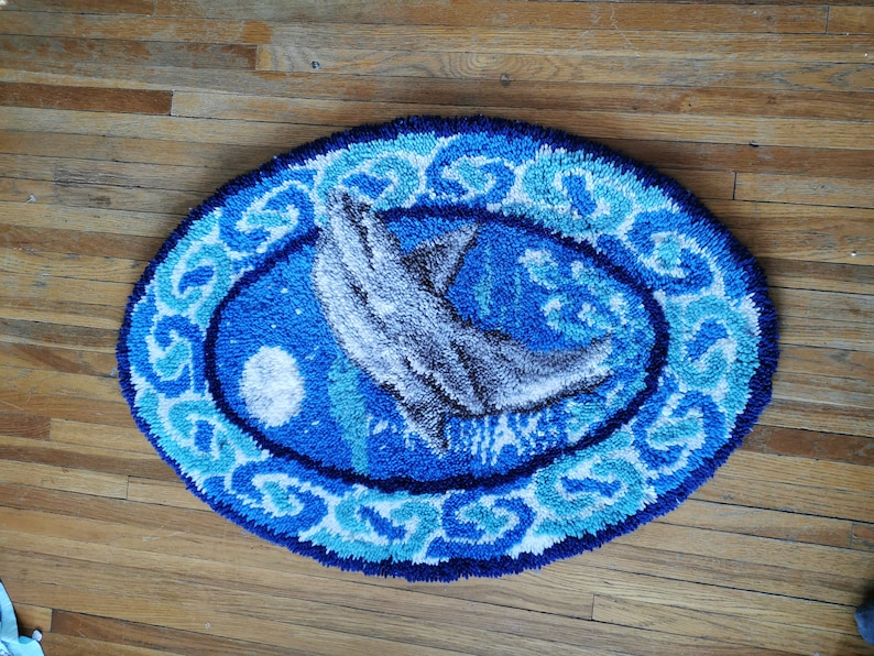 May include: A blue and white oval rug with a white dolphin jumping out of the water. The rug has a blue and white border with a wave pattern.