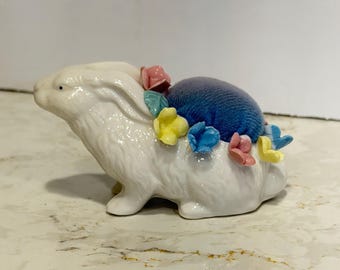 May include: A white ceramic rabbit figurine featuring a dark blue velvet pincushion on its back. The rabbit is decorated with small, colorful ceramic flowers in shades of pink, yellow, and light blue arranged around the base of the cushion.