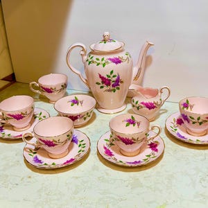 Peut inclure: Un service à thé en porcelaine composé d'une théière avec couvercle, d'un pot à lait, d'un sucrier et de quatre tasses avec soucoupes assorties. L'ensemble présente une base rose pâle ornée de motifs de lilas violets et de feuilles vertes.