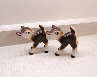 May include: Two brown ceramic deer figurines with white spots. The deer are facing different directions and have pink ears.