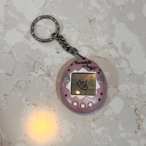 May include: A pink Tamagotchi Angel digital pet with a silver keychain. The device has a small screen displaying a pixelated angel character. The text "Tamagotchi Angel" is printed above the screen.