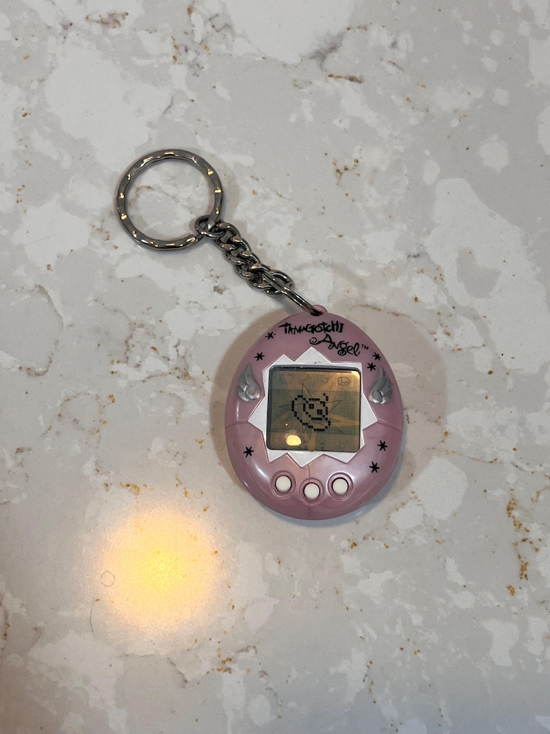 May include: A pink Tamagotchi Angel digital pet with a silver keychain. The device has a small screen displaying a pixelated angel character. The text "Tamagotchi Angel" is printed above the screen.
