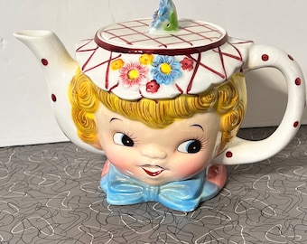 Puede incluir: Una tetera de cerámica blanca con forma de cabeza de niña con una tapa a cuadros rojos y blancos. La tetera tiene un lazo azul y un diseño floral en la tapa.
