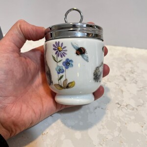 May include: White ceramic egg cup with a silver lid and a floral design featuring a bee, flowers, and a squirrel.