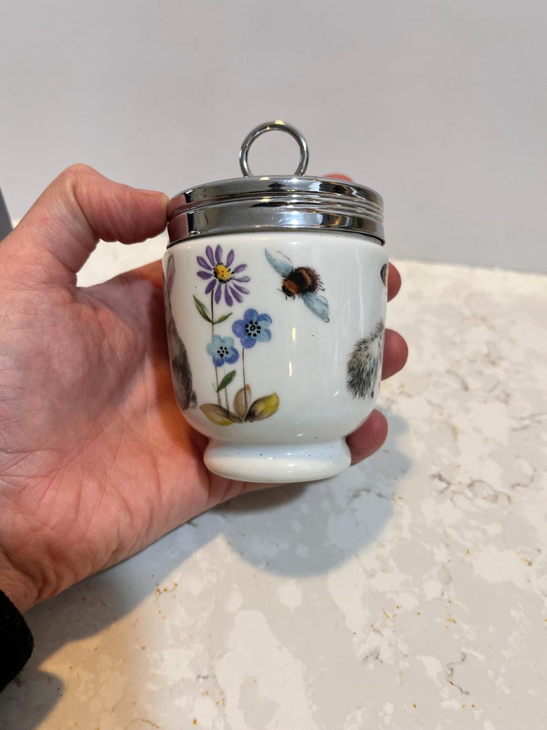 May include: White ceramic egg cup with a silver lid and a floral design featuring a bee, flowers, and a squirrel.