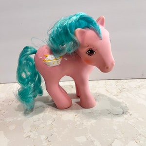 Puede incluir: Un juguete My Little Pony rosa con una melena y cola azules. El pony tiene un símbolo blanco y amarillo en su flanco que representa un tazón con una luna creciente y estrellas.