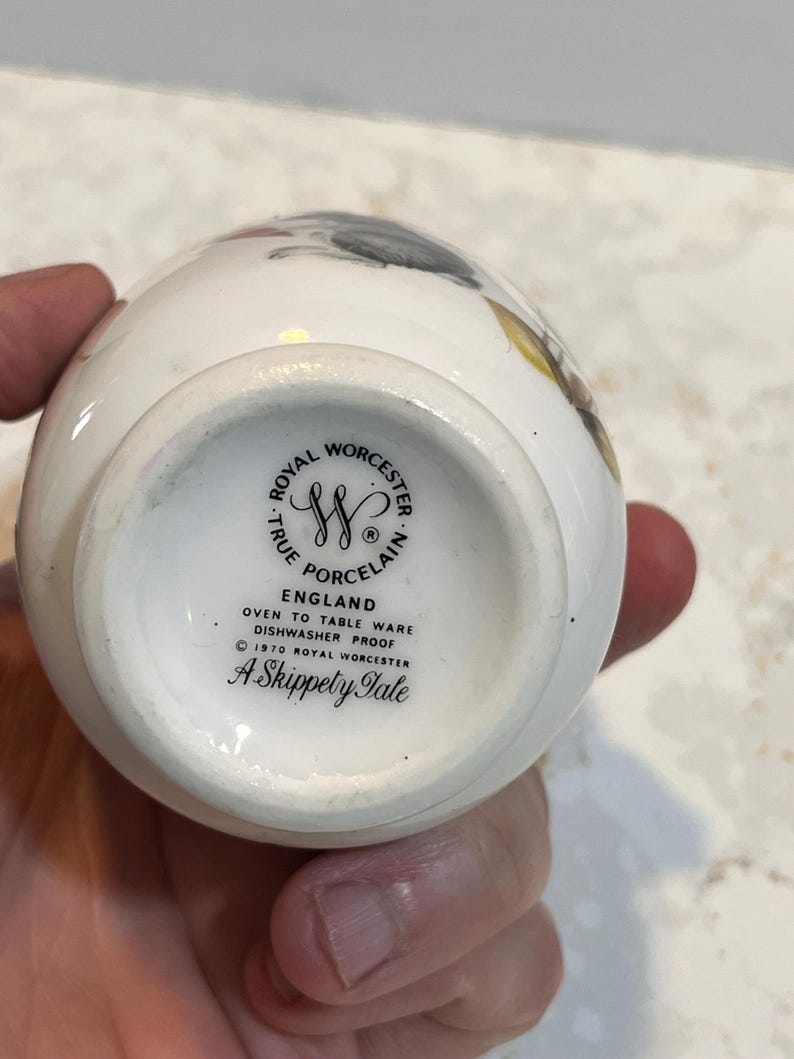 May include: White ceramic egg-shaped vase with a floral design. The bottom of the vase has a black and white logo that reads "Royal Worcester True Porcelain England Oven to Table Ware Dishwasher Proof &copy; 1970 Royal Worcester A Skippety Tale"