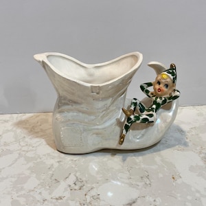 May include: White ceramic planter shaped like a boot with a green and white elf sitting on top. The elf has a yellow face and red lips.