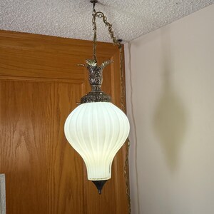 May include: Vintage pendant light with a white, ribbed glass shade. The light fixture has a decorative brass-coloured top and chain. The shade is a teardrop shape. The light hangs from the ceiling in a room with a wooden door.