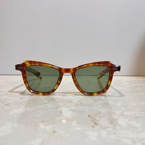 May include: Tortoiseshell-patterned sunglasses with green lenses. The frames have a classic, slightly angular shape. The sunglasses are set against a neutral background, highlighting their design and color.