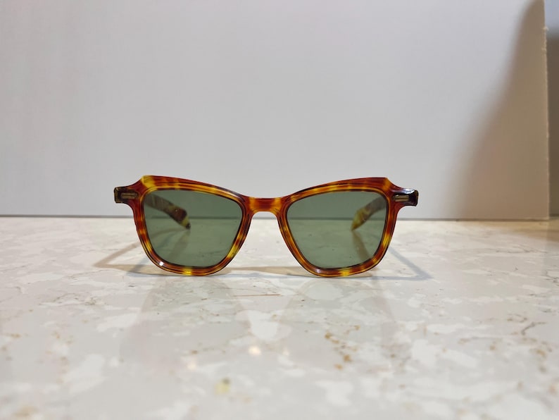 May include: Tortoiseshell-patterned sunglasses with green lenses. The frames have a classic, slightly angular shape. The sunglasses are set against a neutral background, highlighting their design and color.