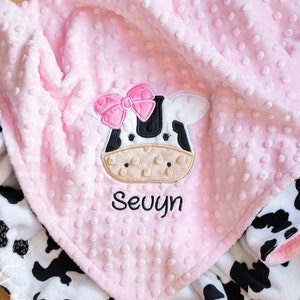 May include: A pink baby blanket with a cow design. The blanket has a pink, textured top layer and a black and white cow print on the underside. The cow face is embroidered with a pink bow and the name "Sevuyn".