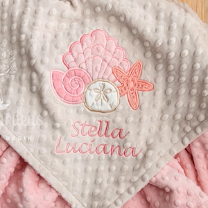 May include: A baby blanket in pink and beige, featuring a seashell and starfish design. The blanket is embroidered with the name "Stella Luciana". The blanket has a textured, dotted surface.