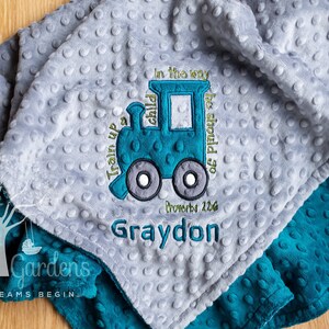 May include: A gray and teal baby blanket with a train design and the text "Train up a child in the way he should go, Proverbs 22:6" and the name "Graydon" embroidered on it.