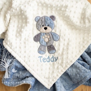 May include: A baby blanket with a white, textured top layer and a blue bottom layer. The blanket features an embroidered teddy bear design in shades of blue and gray, with the name "Teddy" embroidered below. The blanket is folded on a wooden surface.