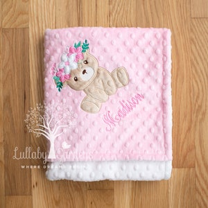 Puede incluir: Una manta rosa para beb&eacute; con un oso de peluche con una corona de flores. La manta tiene una textura punteada y el nombre "Madison" bordado en rosa. El borde inferior es blanco y esponjoso. El logotipo de Lullaby Gardens est&aacute; en la esquina inferior izquierda.