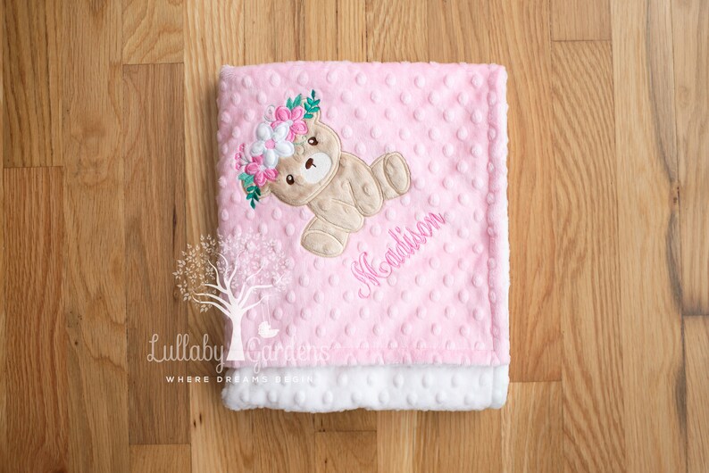 Puede incluir: Una manta rosa para beb&eacute; con un oso de peluche con una corona de flores. La manta tiene una textura punteada y el nombre "Madison" bordado en rosa. El borde inferior es blanco y esponjoso. El logotipo de Lullaby Gardens est&aacute; en la esquina inferior izquierda.