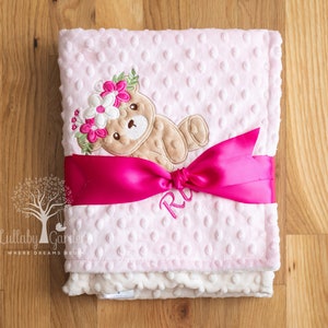 May include: A pink baby blanket with a teddy bear design wearing a floral crown. The blanket is wrapped with a bright pink ribbon and features the embroidered name "R". The blanket is made of soft, textured material.