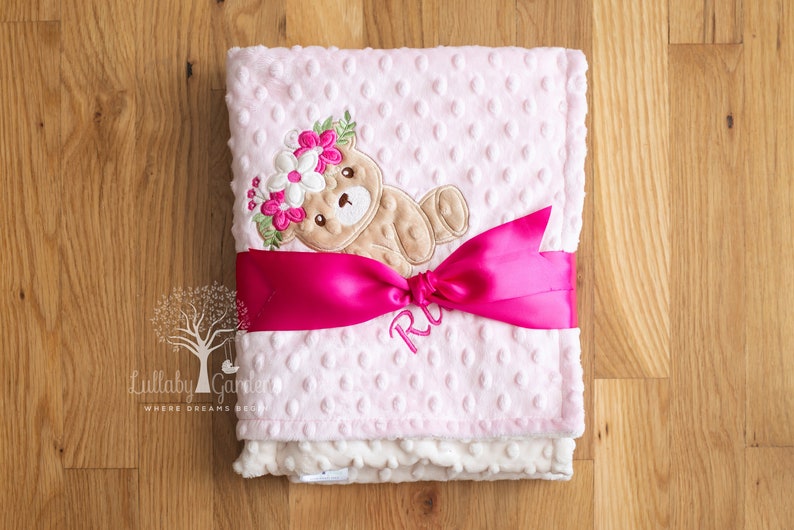 May include: A pink baby blanket with a teddy bear design wearing a floral crown. The blanket is wrapped with a bright pink ribbon and features the embroidered name "R". The blanket is made of soft, textured material.