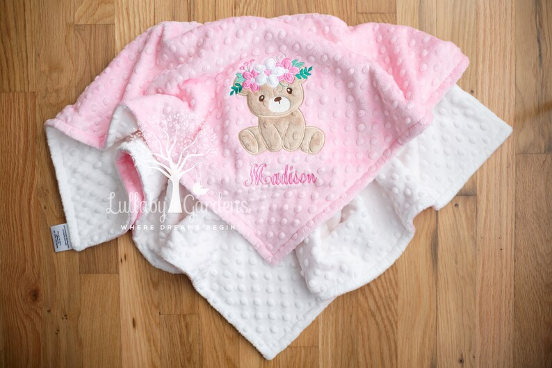 Puede incluir: Una manta rosa y blanca para beb&eacute; con un dise&ntilde;o de oso de peluche. La manta rosa tiene una superficie texturizada y punteada y presenta un oso bordado con una corona floral. El nombre "Madison" est&aacute; bordado debajo.