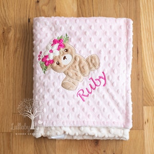 May include: Pink baby blanket with a teddy bear wearing a floral crown. The blanket has a dotted texture and the name "Ruby" embroidered in pink. The blanket is made of soft material.