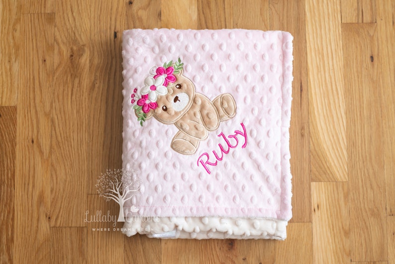 May include: Pink baby blanket with a teddy bear wearing a floral crown. The blanket has a dotted texture and the name "Ruby" embroidered in pink. The blanket is made of soft material.