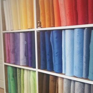 May include: A multi-shelf storage unit filled with rolls of colorful fabric. The fabric rolls are arranged by color, ranging from white, yellow, orange, red, purple, blue, green, brown, and gray. The fabric has a textured, dotted surface.