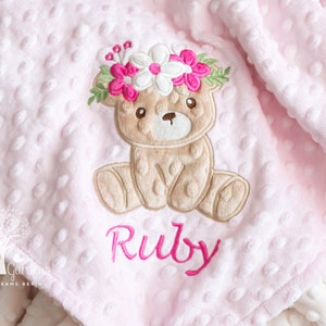 May include: Pink baby blanket with a textured dot pattern. Features an embroidered teddy bear wearing a floral crown, with the name "Ruby" in pink script. The blanket is soft and plush.