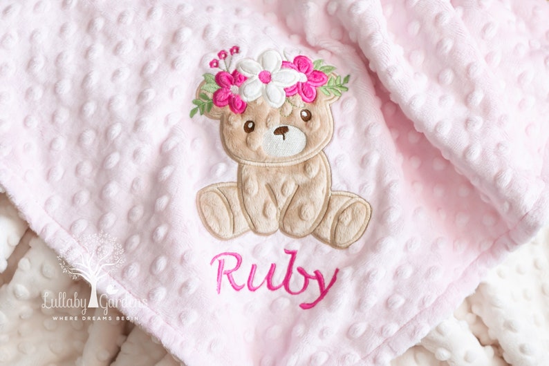 May include: Pink baby blanket with a textured dot pattern. Features an embroidered teddy bear wearing a floral crown, with the name "Ruby" in pink script. The blanket is soft and plush.