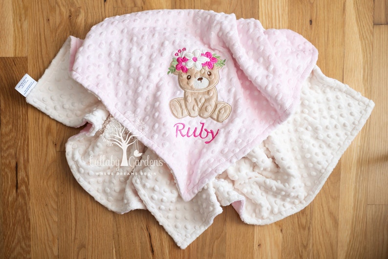 May include: A soft, pink baby blanket with a textured dot pattern. The blanket features an embroidered teddy bear wearing a floral crown and the name "Ruby" in pink script. A second blanket in cream is visible.