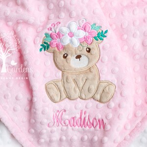 Puede incluir: Manta rosa para beb&eacute; con una superficie texturizada y punteada. Un oso de peluche bordado con una corona de flores est&aacute; centrado en la manta. El nombre "Madison" est&aacute; bordado en rosa debajo. La manta tiene un reverso blanco.