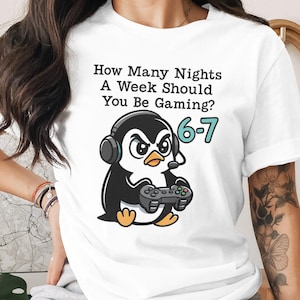 May include: White t-shirt featuring a cartoon penguin wearing a headset and holding a game controller. Text above the penguin reads: "How Many Nights A Week Should You Be Gaming?" with "6-7" in teal. A fun, casual tee.